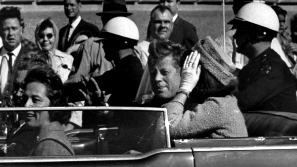 अमेरिका के पूर्व राष्ट्रपति जॉन एफ. कैनेडी 22 नवंबर 1963 को गोली मारे जाने से लगभग एक मिनट पहले अपनी कार के काफिले में हाथ हिलाते हुए।